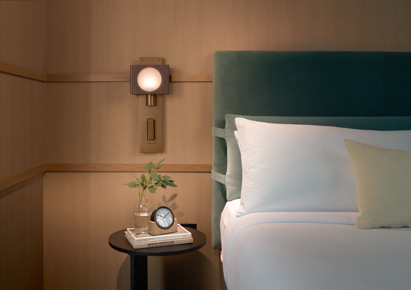Bed with upholstered headboard and luxury linens, on a light woodgrain wall with unique sconce