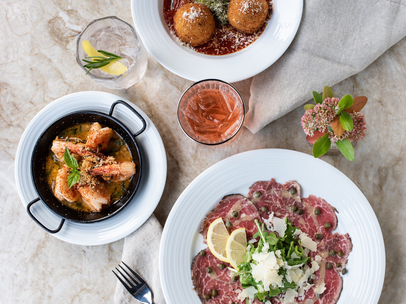 Top-down view of a marble table with shrimp scampi al forno, beef carpaccio and arancini
