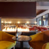 Two gold velvet lounge chairs beside a small black table and bottle of wine, with a fireplace in the background at a restaurant