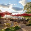 Waterfront patio with black dining tables and chairs, and red umbrellas, with the harbor and willow trees in the background