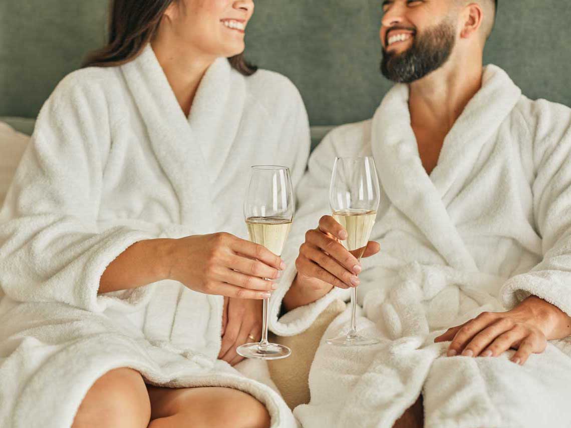 A man and woman wearing white bath robes hold champagne glasses while sitting in bed